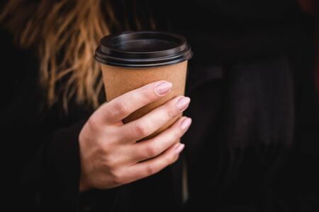 Female hand with paper cup of coffee take away. Woman holding cup on the red wall. Sunny afternoon.の写真素材