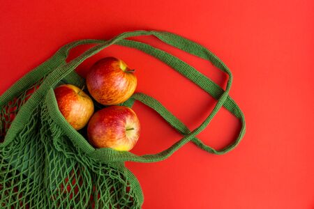 Green reusable shopping string bag with Fresh apples on red background. Zero waste concept. No...