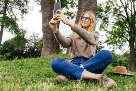 Beautiful young hipster woman taking selfie on green grass at the park. Looking at camera and smile....