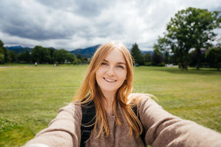 Beautiful young hipster woman taking selfie in the public park. Looking at camera and smile. Travel and active life concept. Outdoorsの写真素材