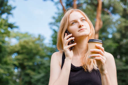 Beautiful young hipster woman with blonde hair rest at the park. Female freelancer drinking coffee...