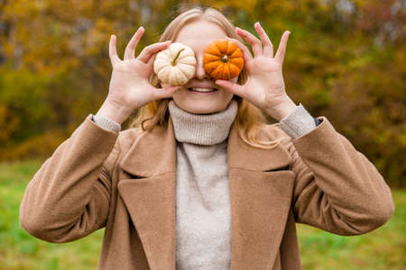 The girl covers part of her face with an orange and white pumpkins. Harvest concept. Copy space. Autumn lifestyle. Smiling Woman being playful covering her eyes with pumpkinsの写真素材