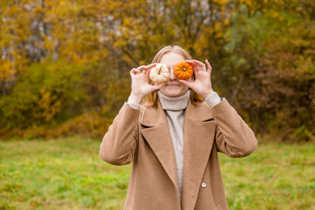 The girl covers part of her face with an orange and white pumpkins. Harvest concept. Copy space. Autumn lifestyle. Smiling Woman being playful covering her eyes with pumpkinsの写真素材