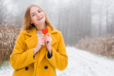 Happy young woman wearing a coat holding a wooden heart over nature background.の写真素材