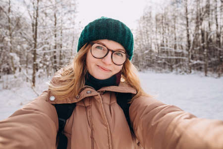 Young beautiful happy girl in green winter knitted hat takes selfie in winter forest. Looking at camera and smile. Travel and active life concept. Outdoorsの写真素材