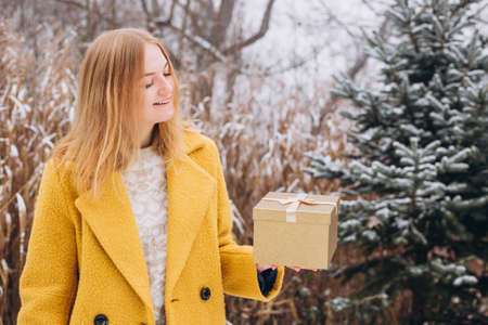 Happy young woman wearing a coat holding a Christmas present box over nature background.の写真素材