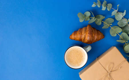 Present box, coffee, croissant and eucalyptus leaves on Blue background. Good Morning, holiday...