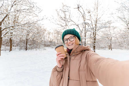 Young beautiful happy girl in winter knitted hat takes selfie in winter forest.