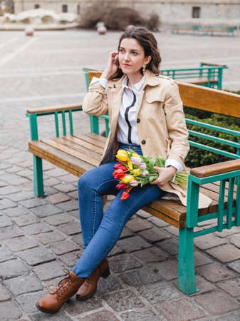 Beautiful young woman with spring tulips flowers bouquet at city street. Happy girl walking outdoors. Spring portrait of pretty female in park.の写真素材