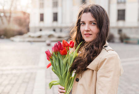 Beautiful young woman with spring tulips flowers bouquet at city street. Happy girl walking outdoors. Spring portrait of pretty female in park.の写真素材