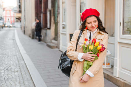 Beautiful young woman with spring tulips flowers bouquet at city street. Happy girl walking outdoors. Spring portrait of pretty female in park.の写真素材