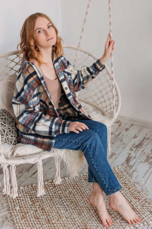 Young blonde woman chilling at home in comfortable hanging chair. Atmospheric hygge styleの写真素材