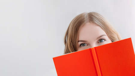 Portrait of a pretty young blond girl hiding behind an open orange book.