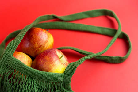 Green reusable shopping string bag with Fresh apples on red background. Zero waste concept. No...