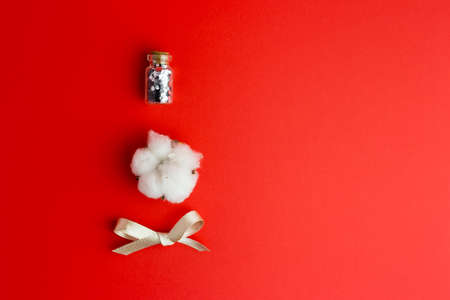 Cotton flower, glass bottle with confetti and bow on red background. Flat lay, Top view with space for text. Holiday or care concept.の写真素材