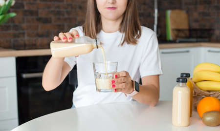 Happy brunette woman sitting with bottle of smoothie and fruits at home kitchen. Vegan meal and detox concept. Girl with white t-shirt drinking fresh cocktail. Mockup of packagingの写真素材