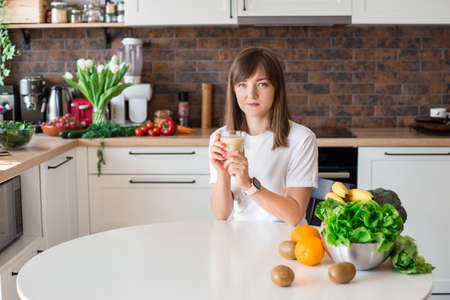 Happy brunette woman sitting with homemade glass smoothie and healthy fruits at home kitchen. Vegan meal and detox concept. Girl with white t-shirt drinking fresh cocktailの写真素材