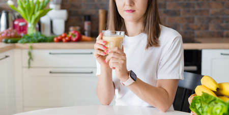 Happy brunette woman sitting with homemade glass smoothie and healthy fruits at home kitchen. Vegan meal and detox concept. Girl with white t-shirt drinking fresh cocktailの写真素材