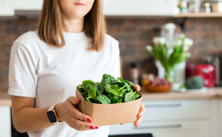 Woman in white t-shirt holding a green spinach in recycling ecology packing in kitchen at home. Raw food diet and healthy eating concept. Baby foodの写真素材