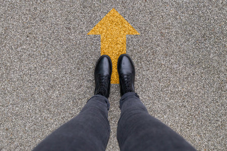 Black shoes standing on the asphalt concrete floor with yellow direction arrow symbol. Moving forward, new start and success.. Feet shoes walking in outdoor. Youth Selphie Modern hipsterの写真素材