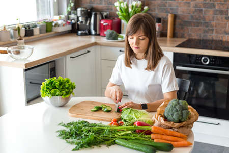 Brunette Woman in white t-shirt preparing vegetable salad in the loft kitchen at home, Wife Cutting ingredients on table. Healthy Food, Vegan Salad. Dieting Concept.の写真素材