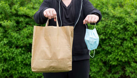 Close up of man hand with mask and paper bag for takeaway food on nature green background. Delivery...