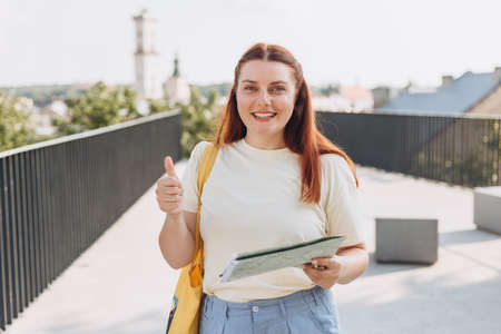 Attractive young female tourist is exploring new city. Happy redhead Woman with map in hands showing thumb up gestureの写真素材