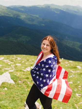 Girl with American Flag looking out at landscape. Young woman stands in the mountains, holding the US flag in her arms high. Flag fluttering in the windの写真素材