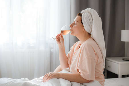 Beautiful happy young woman after shower with glass of wine in bedroom. Staying at home in isolation during quarantine lockdown. Concept of celebrating birthday in bed aloneの写真素材