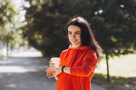 Stylish girl in red sweater looking at the watch and drinking coffee. Happy woman is waiting friends and relaxing in the park, outdoors, summer timeの写真素材