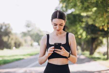 Stylish girl in black top posing on alley holding smartphone. Happy woman with beautiful smile chatting on cell smartphone while relaxing in the park, outdoorsの写真素材