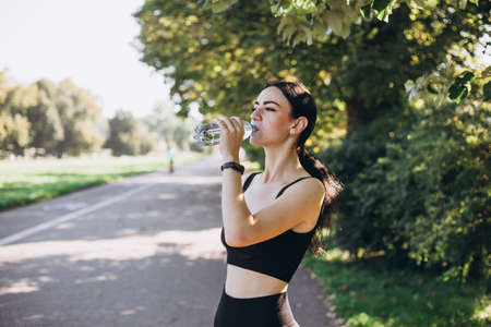 Athletic young woman after runing in the park jogging in the morning for lifestyle health. Female runner standing outdoors holding water bottle. Fitness woman taking a break after running workout.の写真素材
