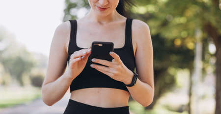 Stylish girl in black top posing on alley holding smartphone. Happy woman with beautiful smile chatting on cell smartphone while relaxing in the park, outdoorsの写真素材