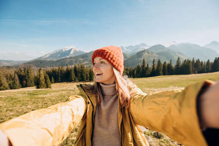 Young woman making selfie photo high in the snow mountains enjoying the view. Freedom, happiness, travel and vacations concept, outdoor activities, she wearing red hat and a yellow jacketの写真素材