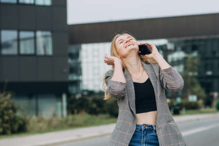 Portrait of young happy blond woman with phone walking on the street in the city. Technology or people concept. Excited girl laughing on bulding background.の写真素材