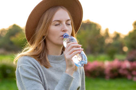Close up portrait of an attractive young blonde woman in hat drinking water from plastic bottle at...