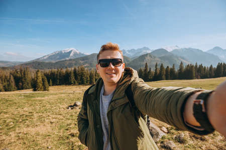 Young man in sunglasses making selfie photo high in the snow mountains enjoying the view. Freedom, happiness, travel and vacations concept, outdoor activities, he wearing a green jacketの写真素材