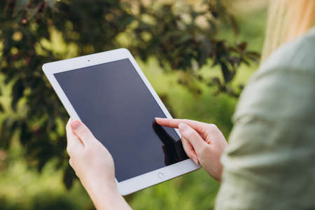 A farmer with a tablet in her hands stands in green garden. Woman smiling, looking at tablet screen. Modern lifestyle and relaxation concepts. Mock upの写真素材