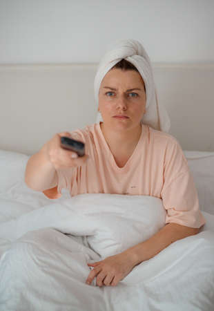 Young unhappy woman lying on bed and watching television. Angry girl with towel sit indoors at home watch tv holding remote controlの写真素材