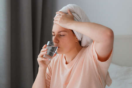 Young woman after shower with glass of water in bedroom. Staying at home in isolation during quarantine lockdown. Woman with headache holding hand on head. new variant - Omicronの写真素材