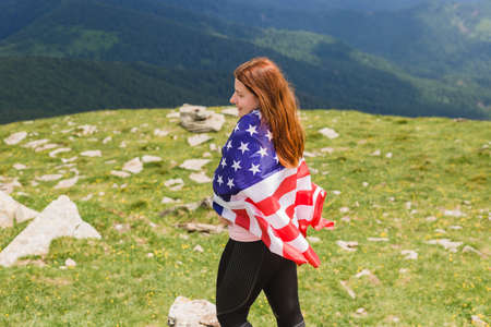 Girl with American Flag looking out at landscape. Young woman stands in the mountains, holding the US flag in her arms high. Flag fluttering in the windの写真素材