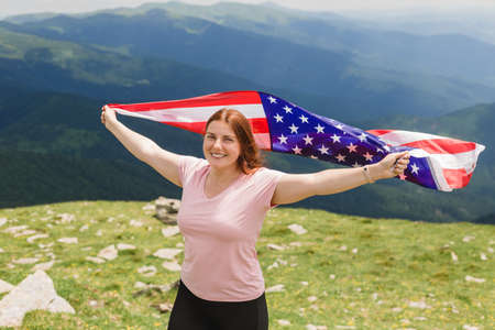 Girl with American Flag looking out at landscape. Young woman stands in the mountains, holding the US flag in her arms high. Flag fluttering in the windの写真素材