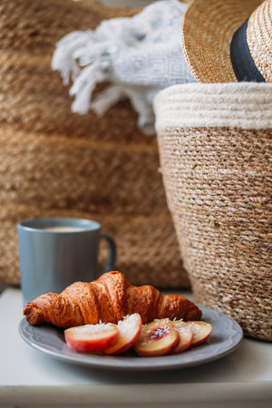 Freshly baked croissant and coffee mug. Coffee in cup on wooden table in cafe. Basket with a gray plaid and a hatの写真素材