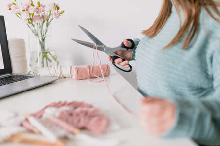 Woman measuring piece of the rope from the coil for macrame weaving with big scissors. Handmade macrame belt in work process. Female hobby.の写真素材