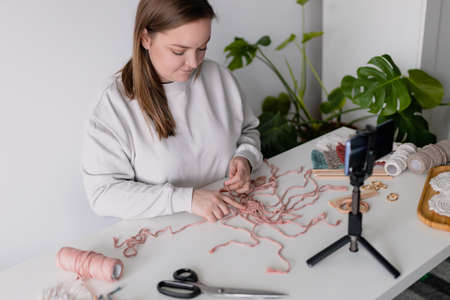 Woman record new videovlog in living room. Puts smartphone on tripod girl make online live stream, lead webinar distant chat, modern tech, video concept. Handmade macrame in work process. Female hobbyの写真素材
