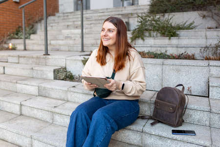 Beautiful smiling female student using tablet outdoors. Happy cheerful young woman siting on stairs on city street, Urban lifestyle concept. Traveler, Online education or shoppingの写真素材