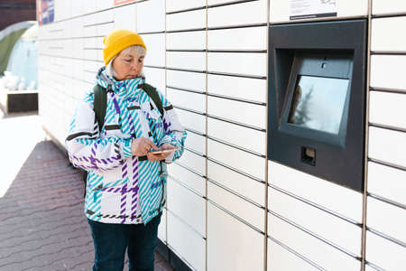 The old woman picked up the package from the parcel locker. Smiling female holding smartphone near...