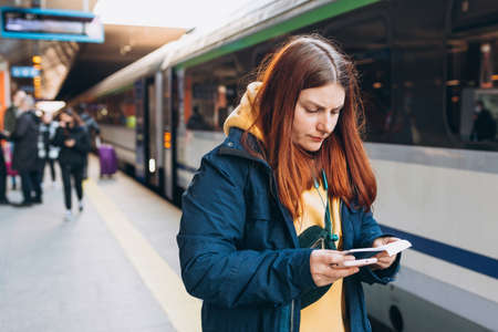 Young redhhead woman with backpack checking her ticket and using smartphone on background train. Railroad transport concept. Girl standing near a platform with blurred railway station.の写真素材