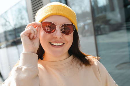 Happy cheerful young woman walking on city street. Portrait of beautiful woman in yellow hat and pink sunglasses smiling outdoors. Urban lifestyle concept. Travelerの写真素材