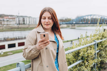 Beautiful happy redhead woman is using a smart phone and smiling outdoors. Urban lifestyle concept. Girl walking on city street checks her smartphone. Spring timeの写真素材